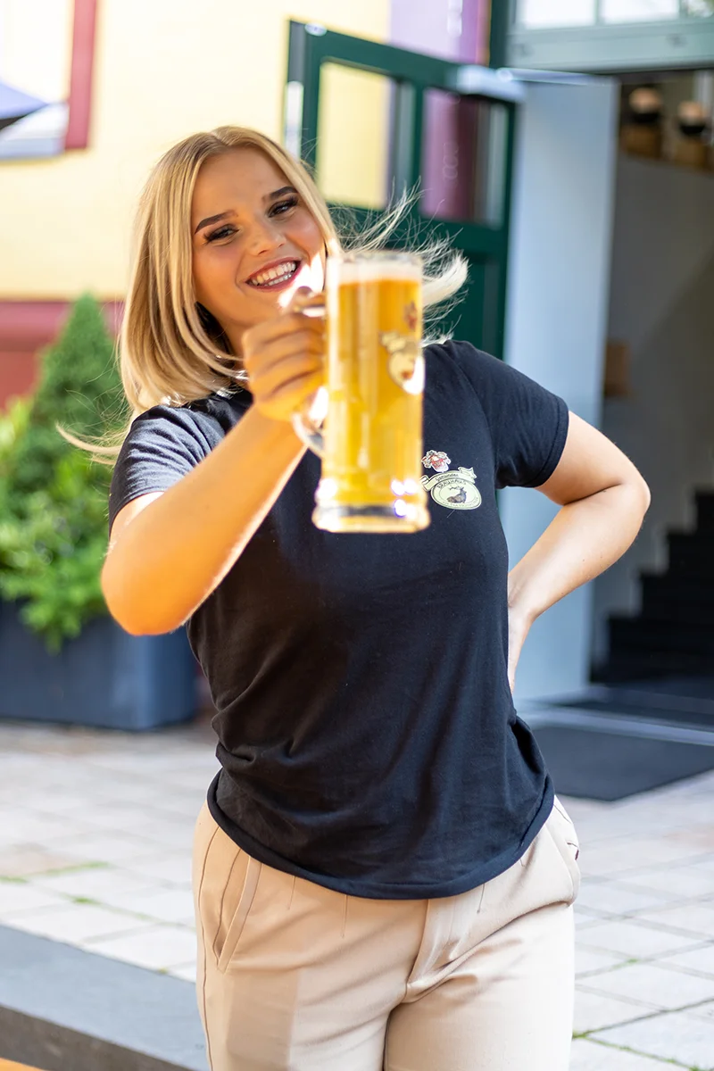 Engagierte Servicekraft des Gemünder Brauhauses strahlt Geselligkeit aus und hebt einen Bierkrug mit geschmackvollem Eifeler Landbier im heimischen Biergarten hoch.